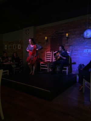 A girl dance on a stage in a small bar along with a man sitting on a chair playing guitar.